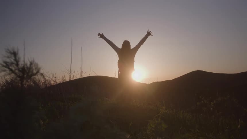 A person stands on a hill with their arms raised and the sun visible behind them