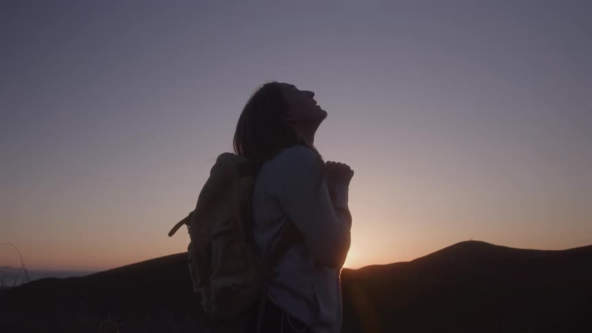 A silhouetted woman with a backpack stands on a mountain and looks up at the sky with the sun rising over the hills in the background