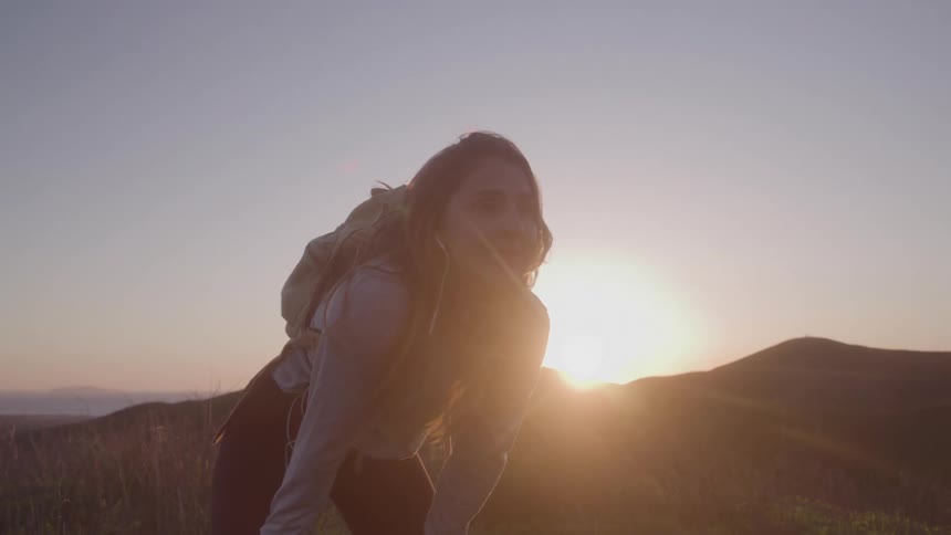 A woman with a backpack and a hoodie is on a hill with other hills in the distance
