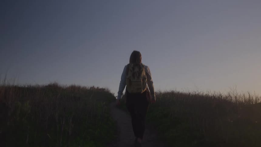 Person walking on a trail at sunset wearing a backpack and lit by the soft light of the sky