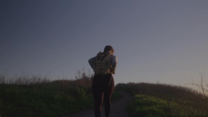 Young woman hiking up a grassy hill in the wilderness at midday carrying a backpack under bright sunlight with open skies above and natural terrain stretching around her