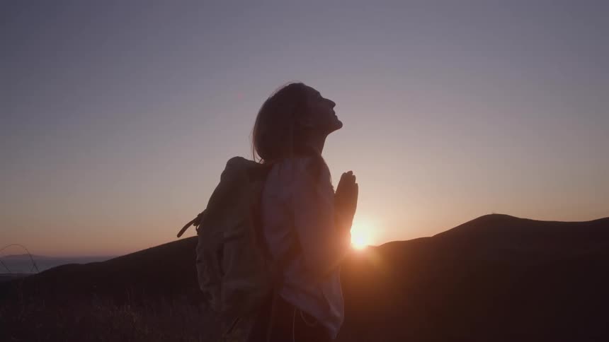 A young woman with a backpack silhouetted against a mountain sunset closes her eyes and clasps her hands in prayer or contemplation