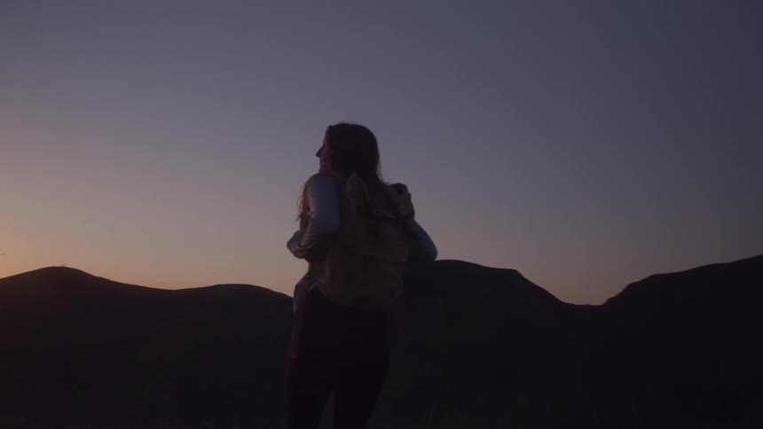 A silhouetted hiker with a backpack walks then stops on a mountain ridge at sunset admiring the view