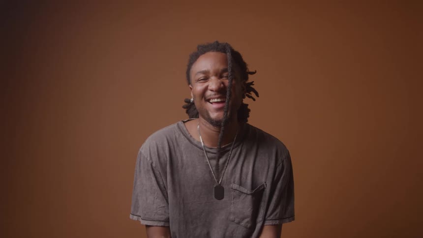 A young man sits and laughs in an indoor setting with a brown background