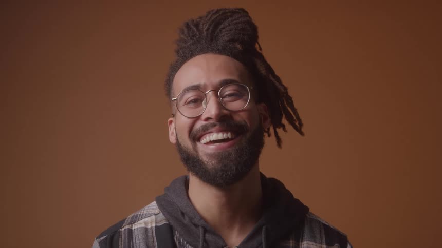 A young black man with thick braided hair and a bearded face stands in front of a brown wall wearing clear glasses and laughing warmly