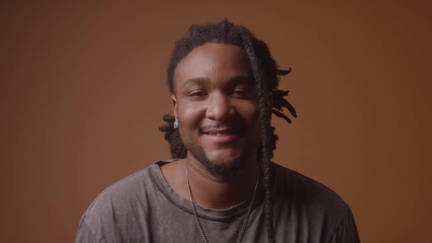 Black male with long thick black braided hair wearing a grey shirt and silver necklace seated in front of a brown wall he has silver earrings and is smiling warmly