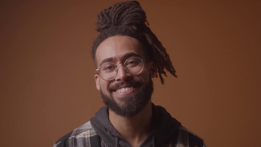 A smiling man with a beard and dreadlocks styled in a top bun wears round glasses and a plaid jacket against a brown background
