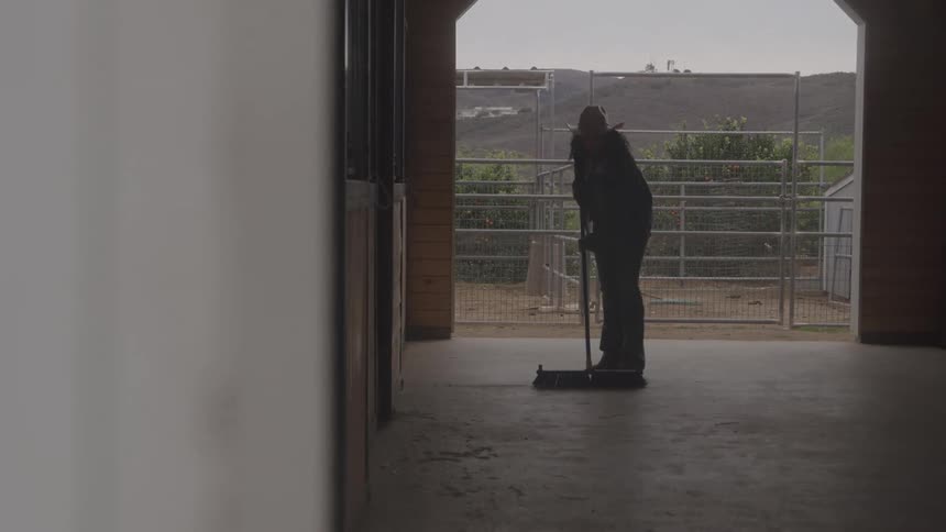 A person in a cowboy hat sweeps the concrete floor of a stable with horse stalls visible on the left and an outdoor corral in the background