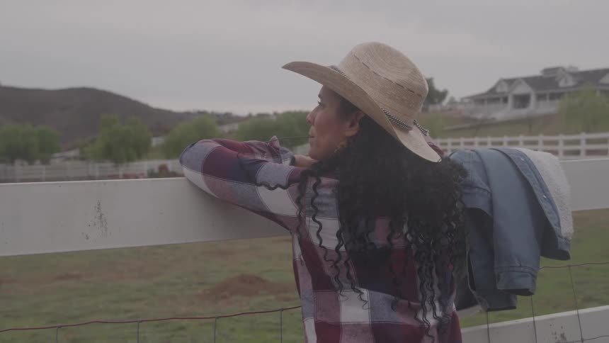 A woman in a cowboy hat and plaid shirt leans on a white fence gazing at the distant hills and a house in the countryside