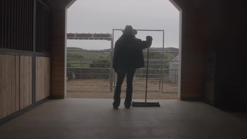 Person wearing a cowboy hat holds a broom in front of a barn gate