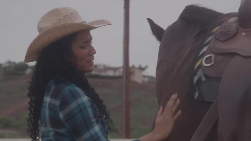 Woman wearing a cowboy hat pets a saddled horse