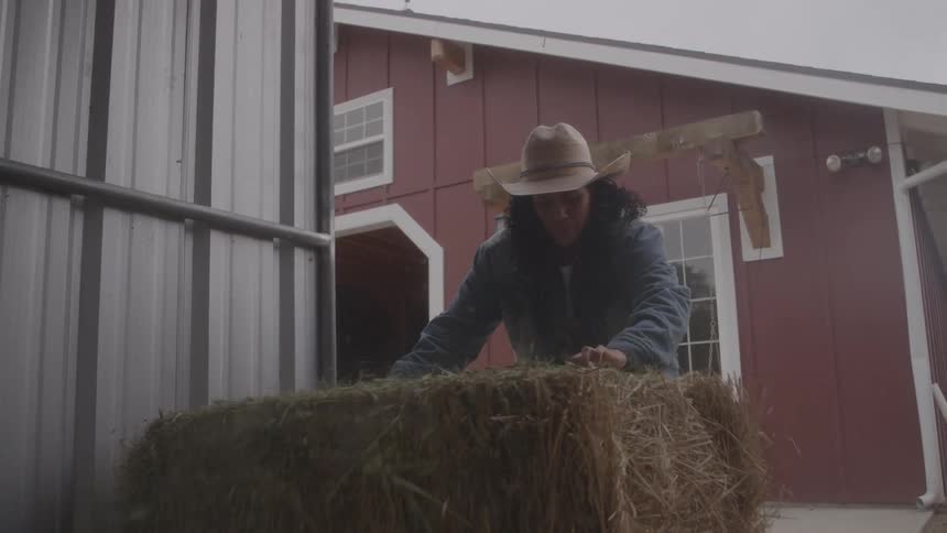 Woman wearing a hat and jeans positions a bale of hay on top of another in a rural area with a red barn in the background