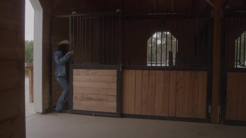 Woman with cowboy hat opens the door of a wooden stable