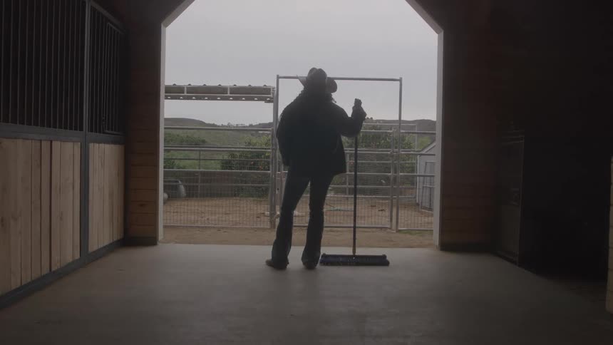 Farmer inside a stable at midday holding a broom wearing a sunhat jean trousers and a jacket standing at the entrance and staring out toward the ranch