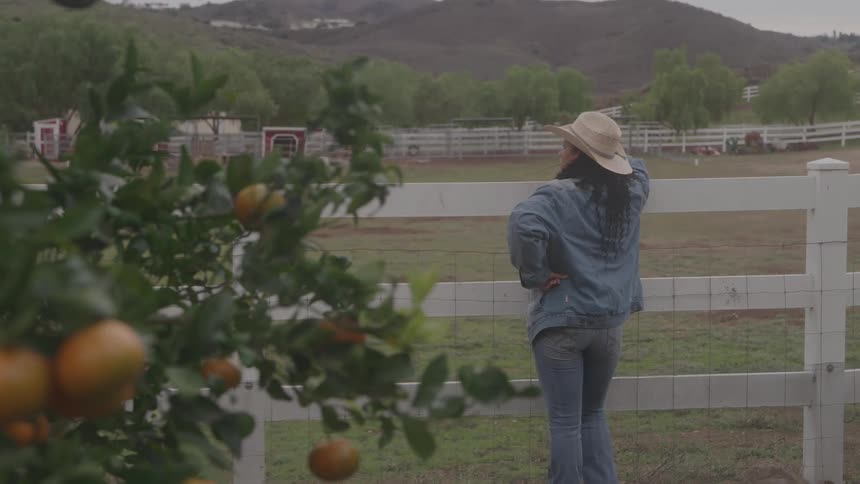 A hispanic woman stands by a fence and looks out at a field