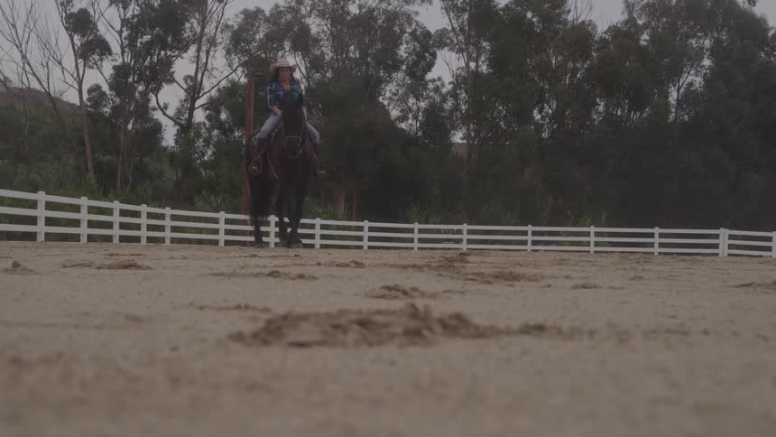 A young woman wearing a cowboy hat rides a horse across a sunlit ranch at midday