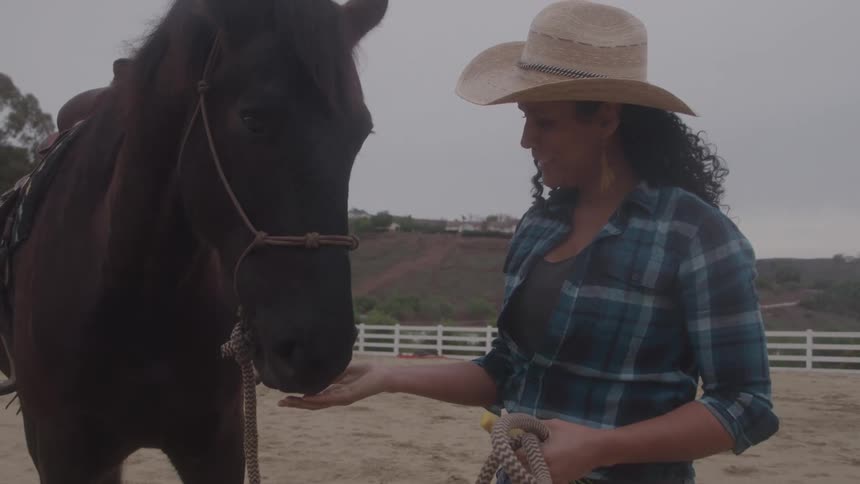 Woman in a cowboy hat feeds a brown horse by hand in a sandy enclosure