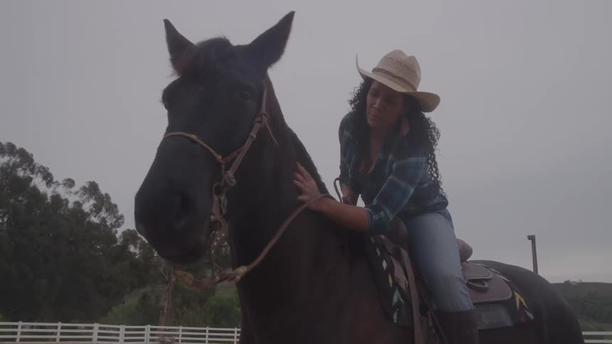 A young woman petting and rubbing her horse while on its back outdoors in a paddock during daytime