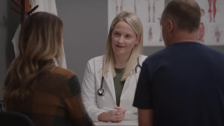 A smiling doctor in a white coat with a stethoscope is talking to a man and a woman in an examination room with a poster of the human nervous system in the background