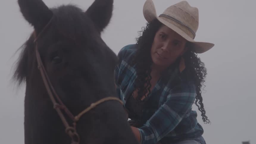 The video shows a closeup of a person with long curly dark hair and a cowboy hat they are sitting on a horse and are gently petting and stroking its head the person appears to be speaking to the horse in a calm and affectionate manner but no audio is