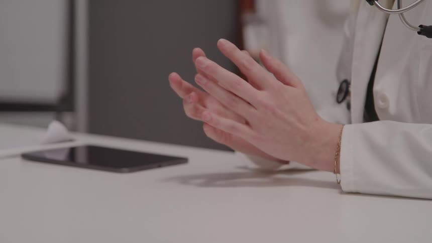 Doctor wearing a white coat and stethoscope keeps hands clasped on the table