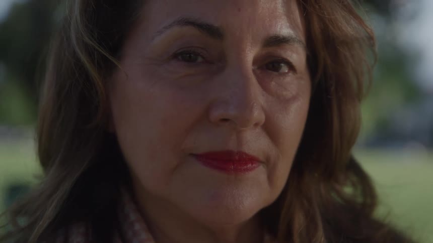 Female face in focus with red lipstick and loose brown hair in outdoor setting with blurred greenery in the background