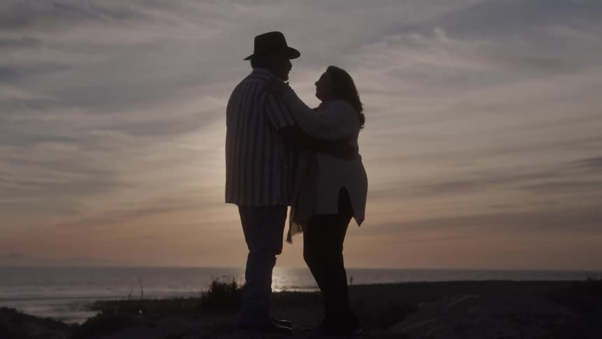 A silhouetted mature man wearing a hat and a striped shirt stands embracing a woman on a hill overlooking the ocean during sunset