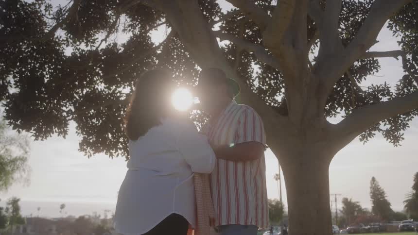 A couple stand facing each other under a tree in an outdoor area