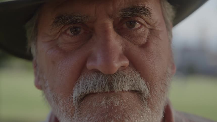 An elderly man with a grey beard wearing a hat standing outdoors with a somber expression