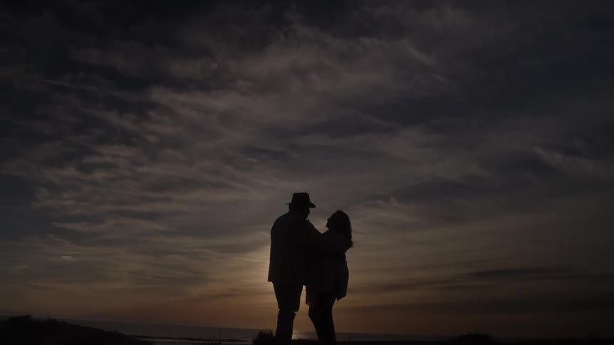 A man and a woman stand and embrace on a hill with the sun and the ocean behind them