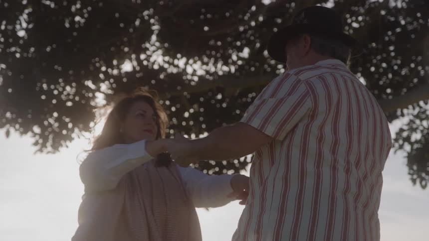 Two people dance under a large tree with sunlight filtering through the branches in a natural setting