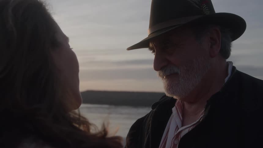 Two people stand close facing and looking at each other outdoors on an overcast beach with a visible water line and horizon during the sunset