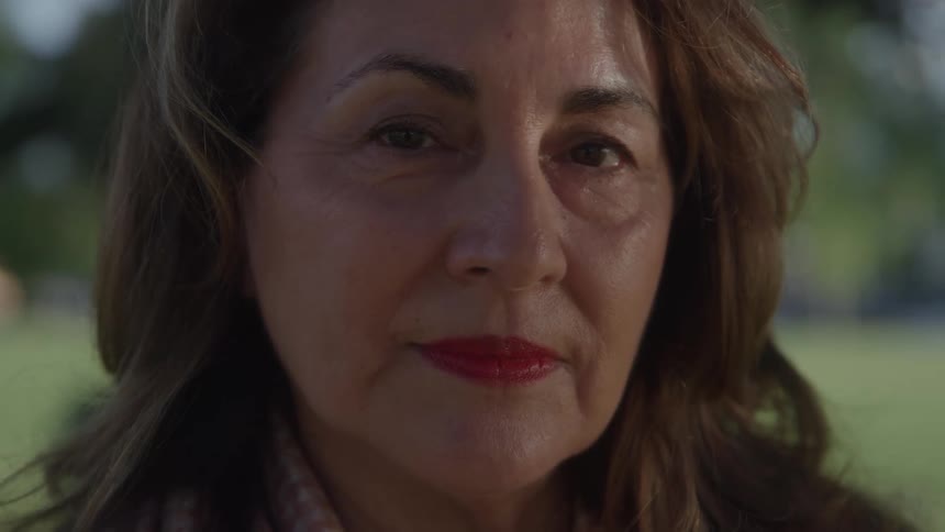 A close view shows a mature woman with long brown hair and red lipstick looking directly at the camera in a park with green grass in the background