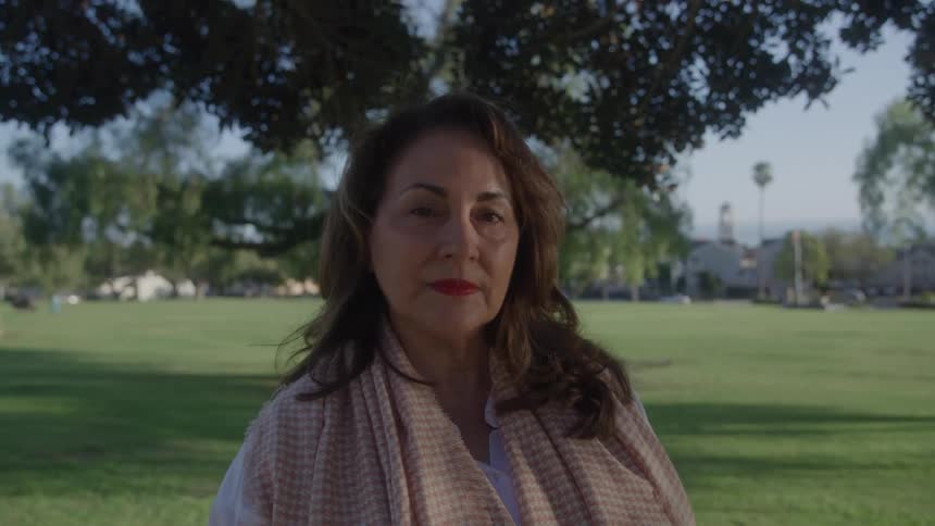 A mature woman with a scarf and a neutral expression looking at the camera on a park setting