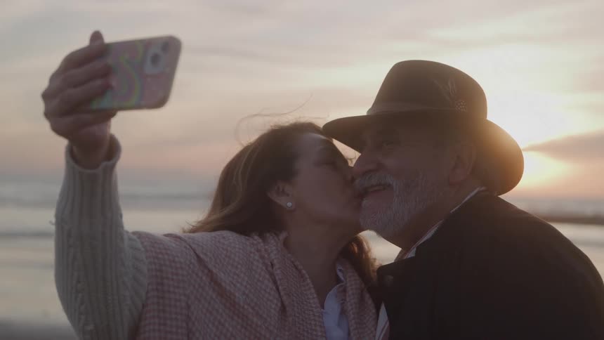 Woman kisses mans cheek while taking a selfie at sunset