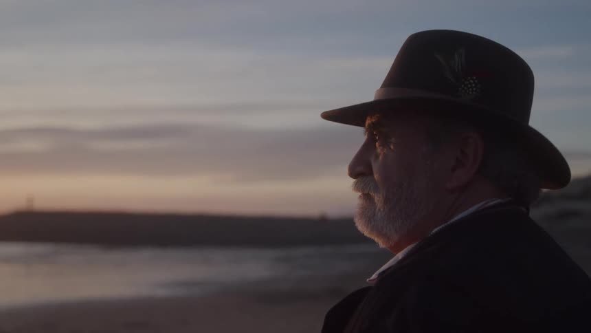 A person with a mustache and a hat stands and looks out at the water during a sunset