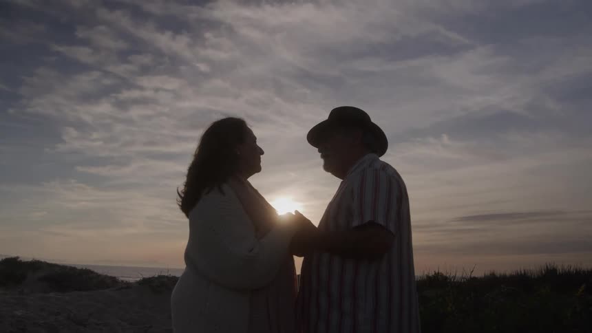 A man and a woman stand on a hill and face each other as they hold hands with the sun setting behind them