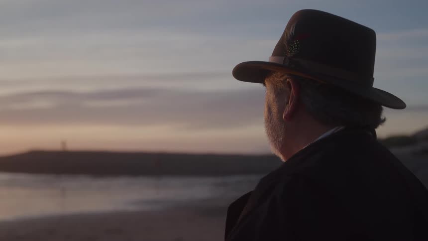 An older man in a brimmed hat watches the shoreline at dusk his steady gaze and slow posture feeling calm and reflective