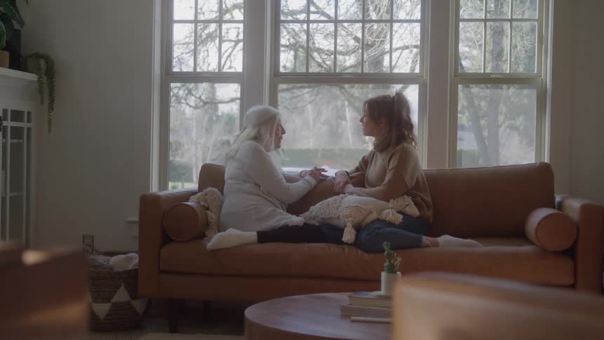 Two women sitting on the couch talking in front of the window