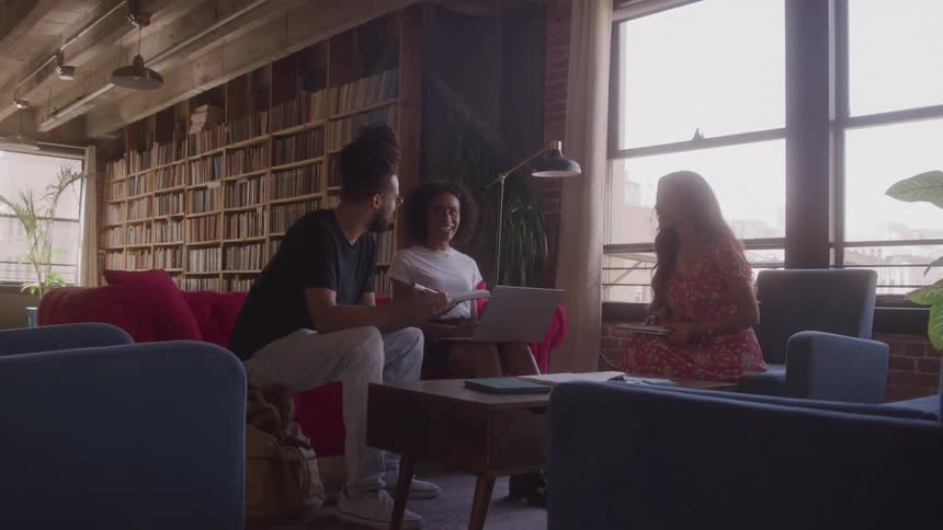 Three people sit in a room with a large bookshelf and have a conversation using laptop and notepads