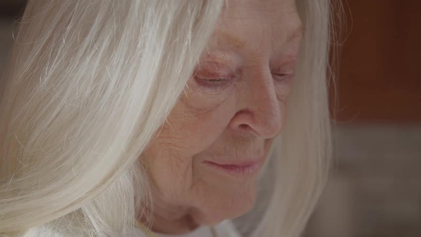 An elderly woman with grey hair sitting indoors at midday softly lit by natural light streaming through a nearby window