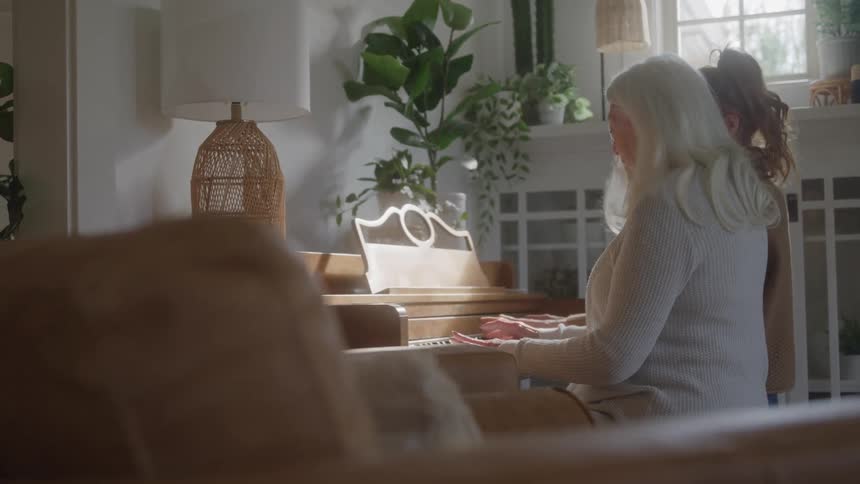 An elderly woman with long white hair sits at a wooden piano with her back to the camera playing the keys while a younger woman stands next to her in a bright plantfilled room