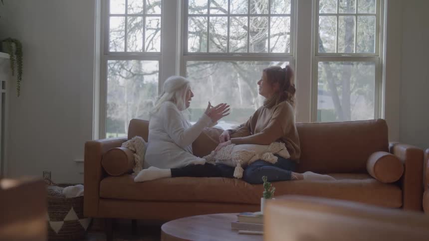 Elderly woman with white hair talks to woman with tiedback hair on brown couch in room lit by large windows with cozy decor