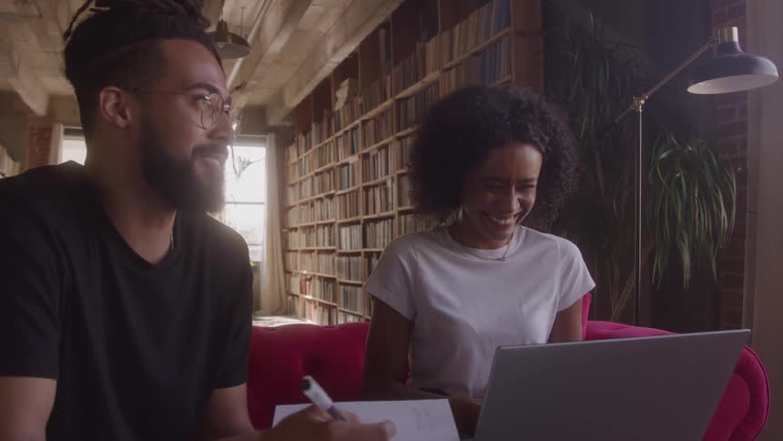 Two young adults sitting inside a library at midday the young woman holds a laptop while the young man clutches a piece of paper both are laughing together clearly amused by something funny theyve discovered surrounded by shelves of books