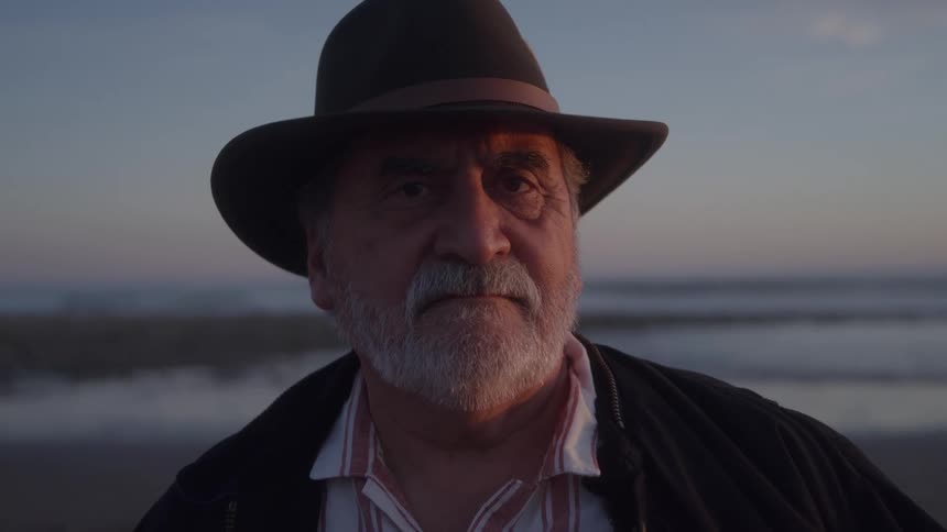 A mature man with a white beard and a fedora hat gazes intently at the camera on a beach at dusk