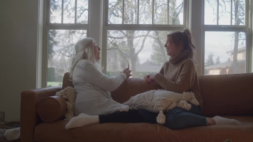 Two women sit on a couch in a room with a large window they lean toward each other talk and laugh