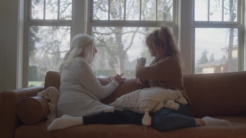 Two women sit on a couch in a room with a window behind them they face each other and talk