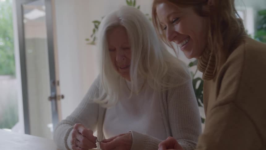An elderly woman with long white hair and a younger woman with reddish hair are sitting at a table together smiling and working on a jigsaw puzzle in a bright modern room