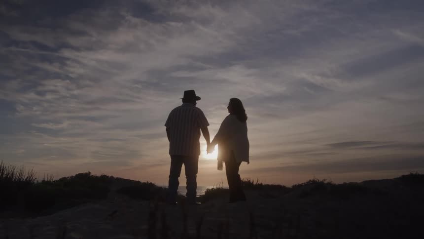 A middleaged couple standing on the beach sand at sunset holding hands as the man wears a hat