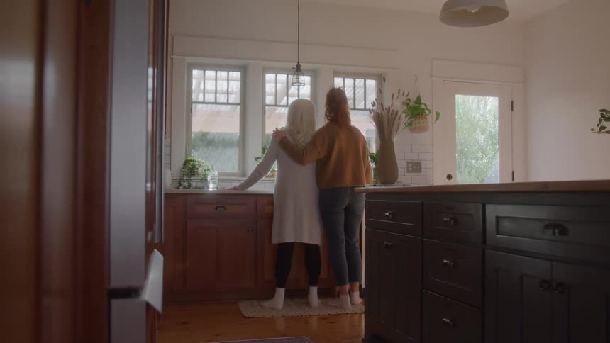 Two women stand side by side in the kitchen looking out the window